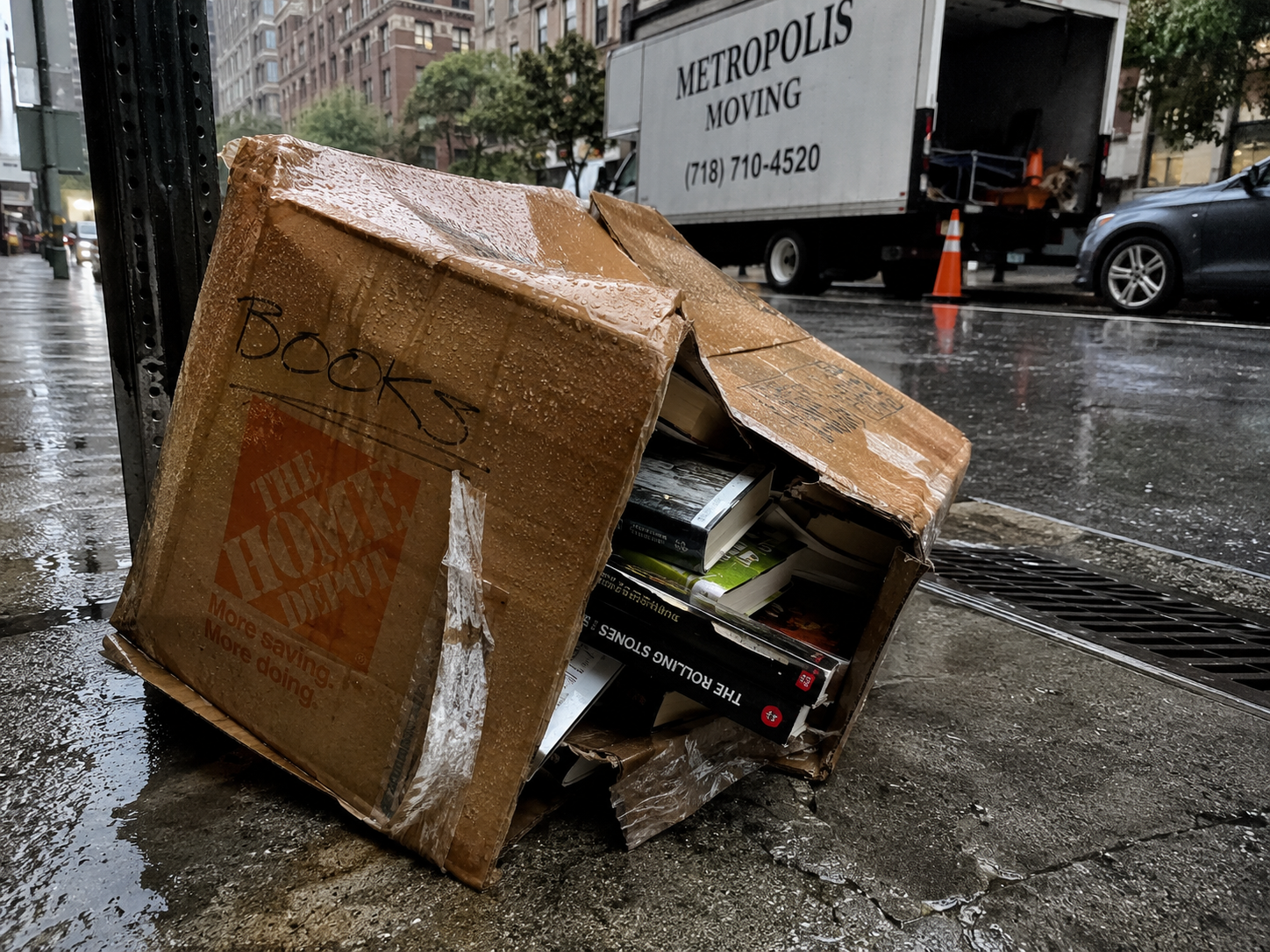 Collapsed damp cardboard moving box on a New York City sidewalk on a rainy moving day with books spilling out and tape peeling off the bottom